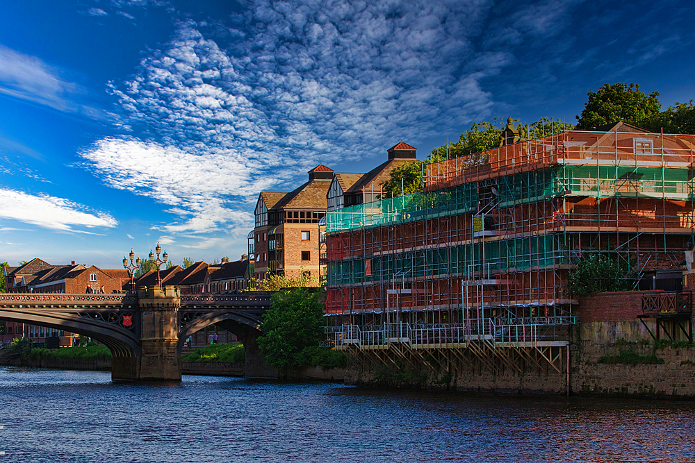 A scenic view of a river with a bridge and buildings under construction on the riverside. The sky is partly cloudy, and the water is calm.