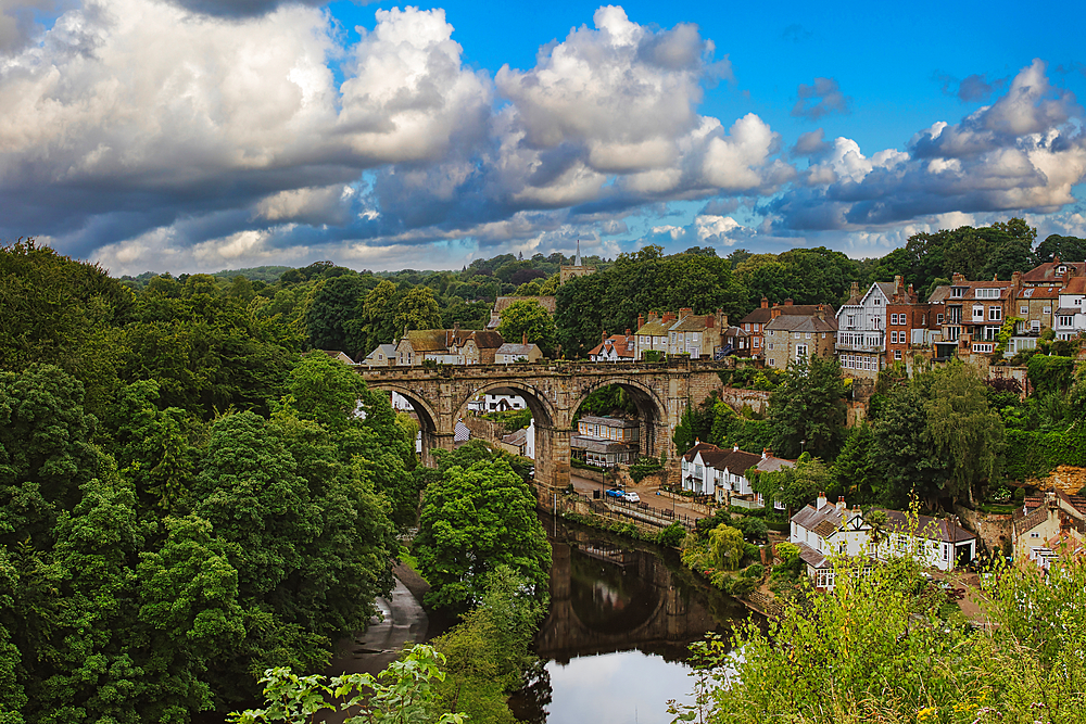 A scenic view of a historic stone bridge over a river, surrounded by lush greenery and quaint houses under a partly cloudy sky.