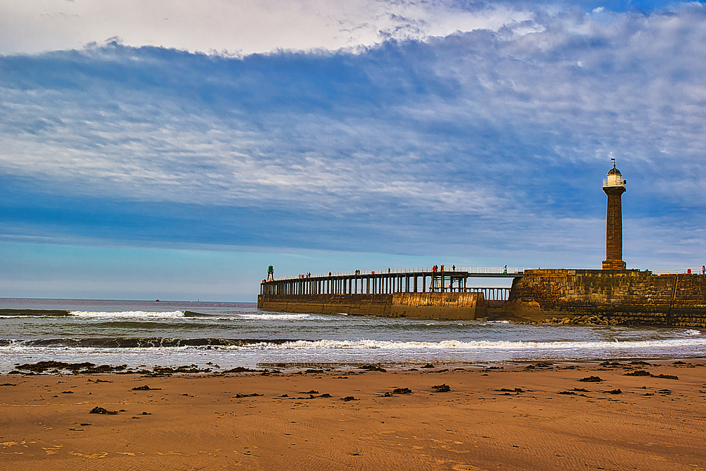 A scenic view of a lighthouse on a pier extending into the ocean, with waves crashing against the shore and a cloudy sky overhead.