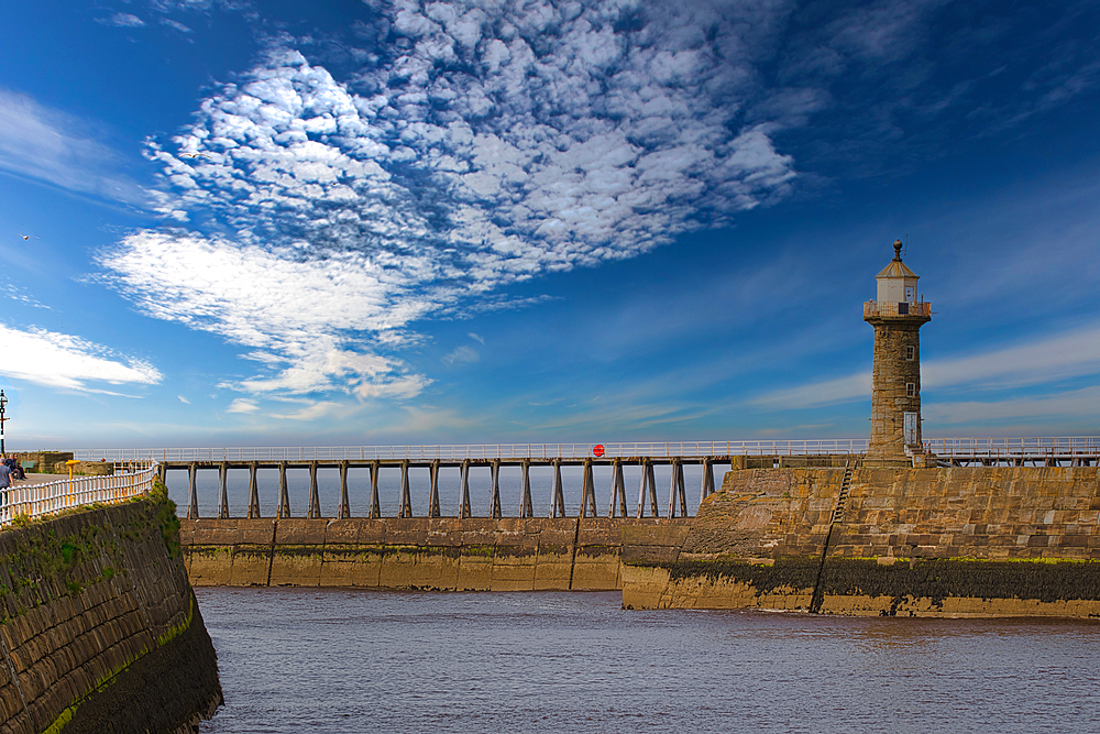 A lighthouse on a stone pier with a cloudy sky in the background. The pier extends into the sea, and the water is calm.