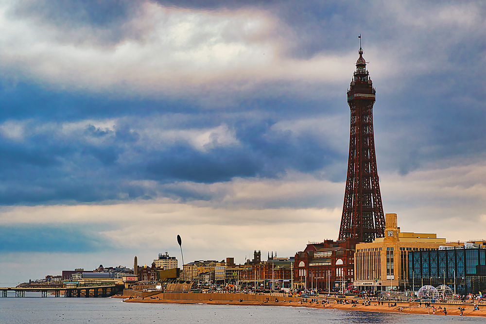 A scenic view of Blackpool, featuring the iconic Blackpool Tower against a dramatic sky. The beach is visible in the foreground with people enjoying the seaside, and the promenade lined with buildings.