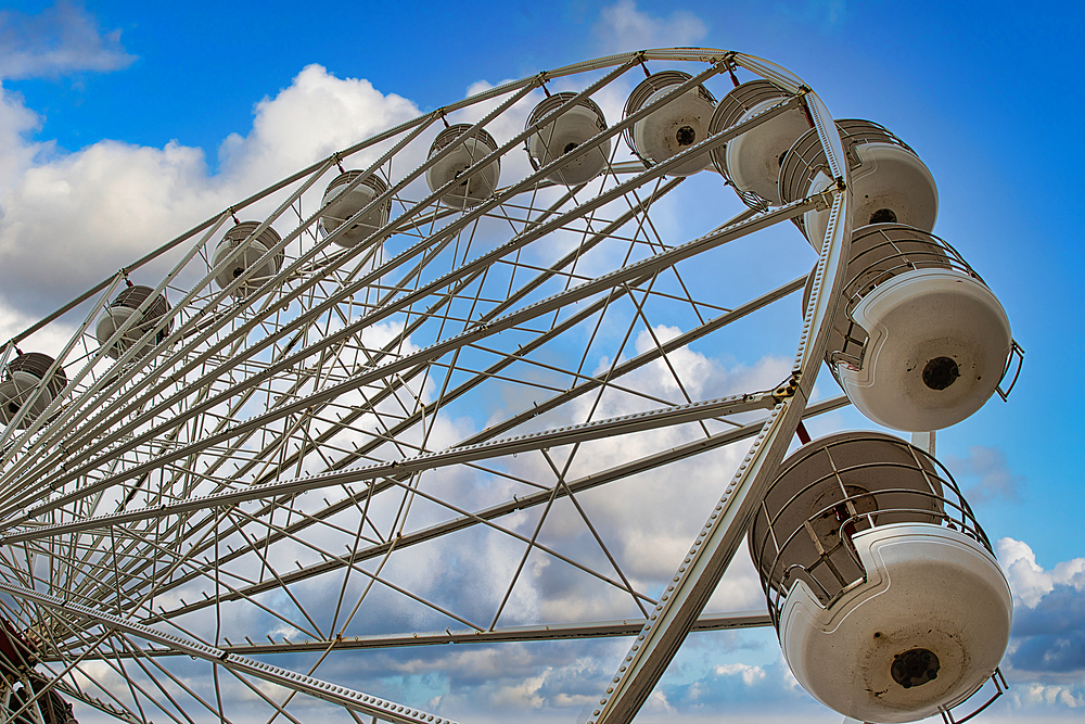 A close-up view of a Ferris wheel against a blue sky with fluffy clouds. The image captures the structure and cabins of the Ferris wheel, showcasing its design and engineering.