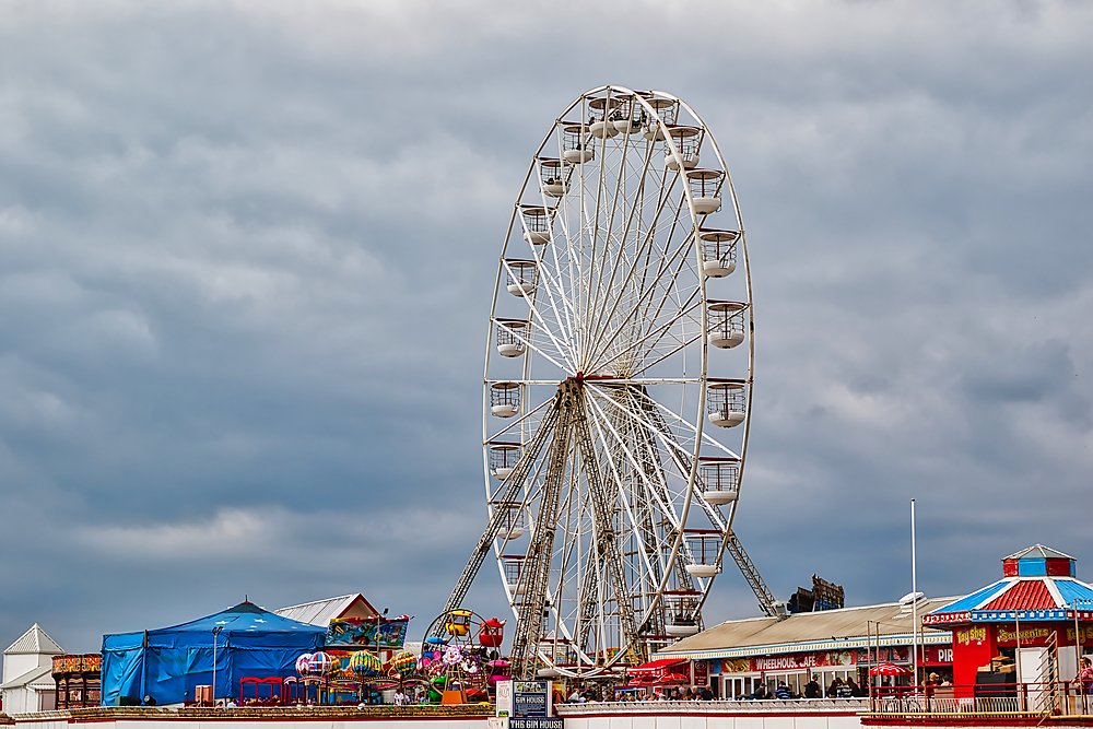 A large ferris wheel stands against a cloudy sky, surrounded by amusement park attractions and colorful structures. The scene captures a lively atmosphere with rides and entertainment.