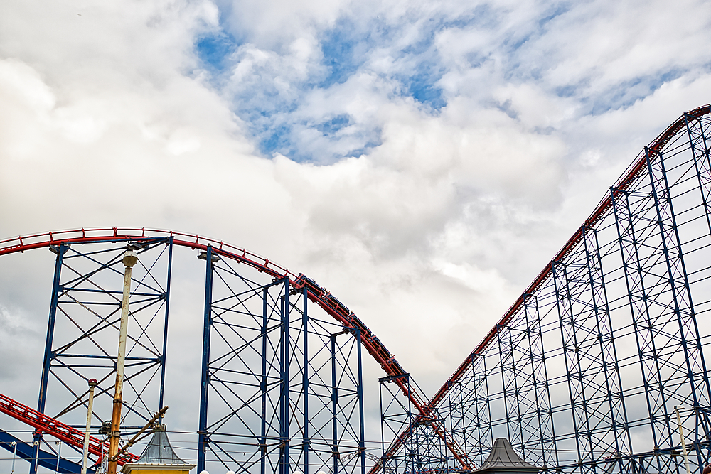 A vibrant roller coaster with red tracks and a steel frame against a cloudy sky. The structure showcases the thrilling design of amusement park rides, emphasizing excitement and adventure.