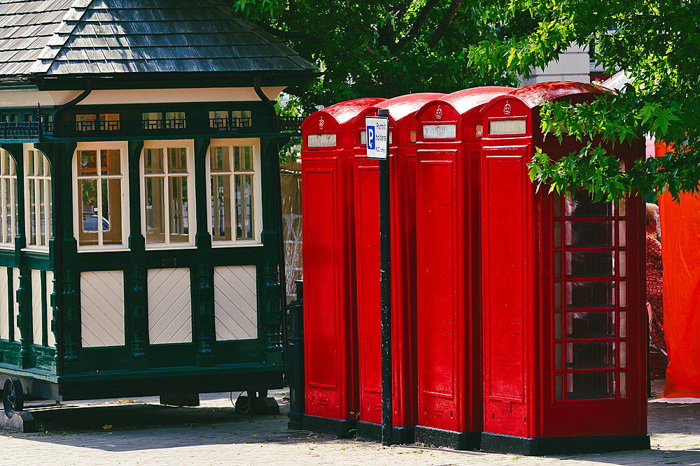 A charming scene featuring three iconic red telephone booths next to a vintage green kiosk. The booths are bright and well-maintained, surrounded by lush greenery and a hint of urban life.