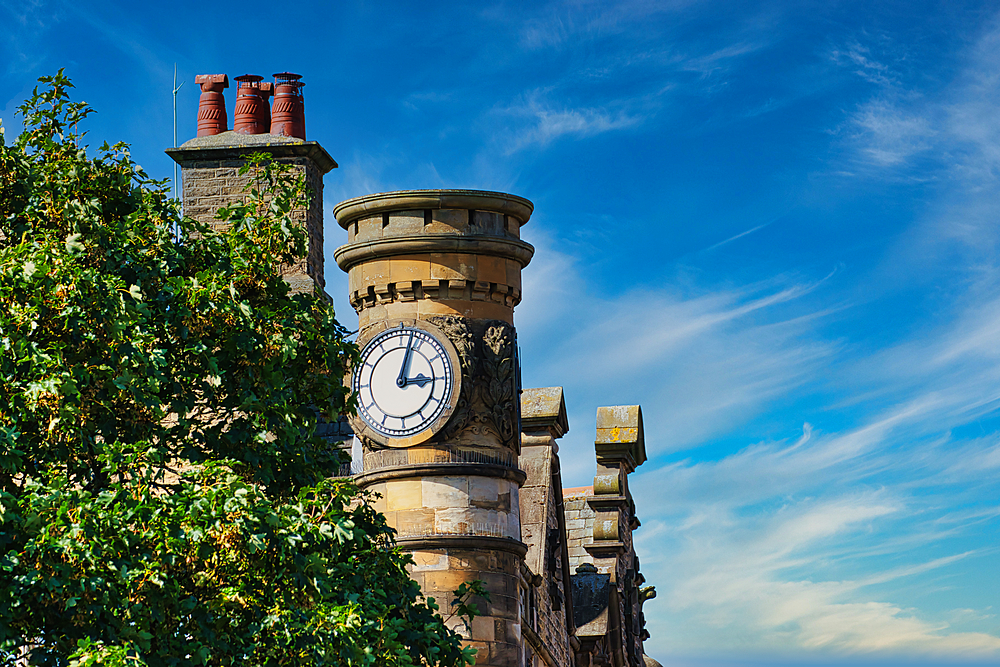 A historic clock tower with a large clock face, surrounded by lush green trees. The sky is bright blue with wispy clouds, creating a picturesque scene.