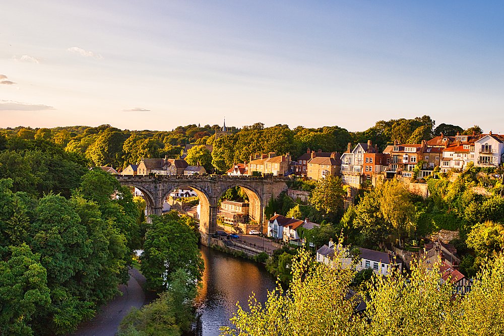 A picturesque view of a stone bridge arching over a river, surrounded by lush greenery and charming houses on the hillside. The scene is illuminated by warm sunset light, creating a serene atmosphere.