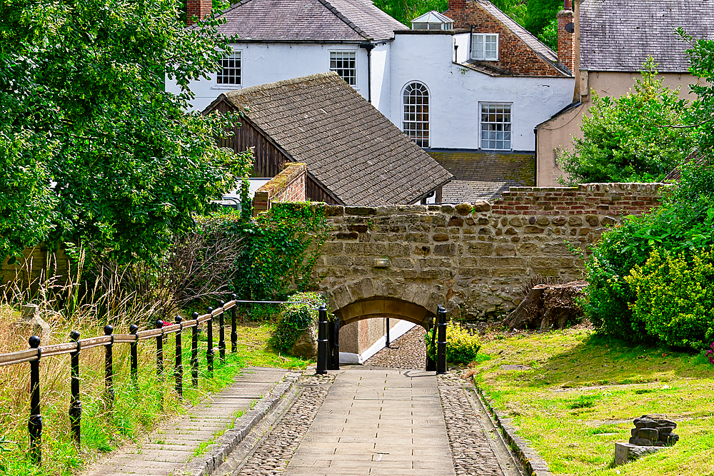 A picturesque pathway leading to a stone arch bridge, surrounded by lush greenery and quaint buildings in the background. The scene captures a serene and charming atmosphere.