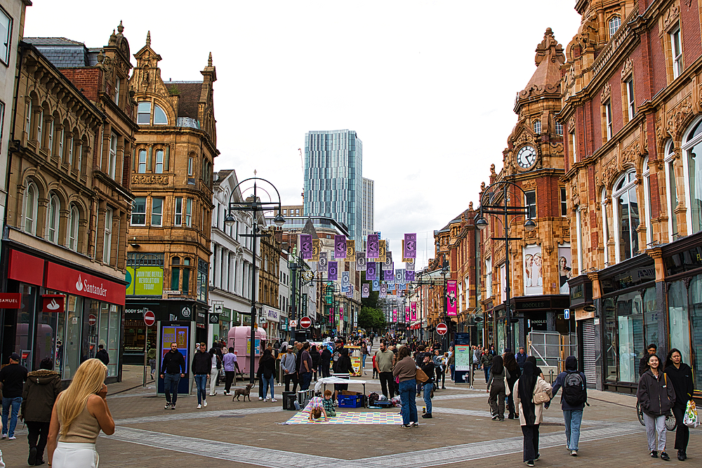 A bustling urban street scene featuring historic architecture, shops, and pedestrians. The street is lively with people walking, and colorful banners hang overhead. Modern buildings are visible in the background, contrasting with the ornate facades of the