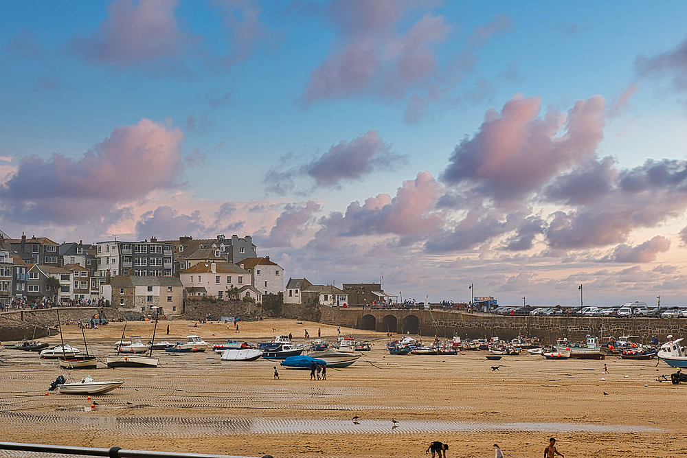 A picturesque coastal scene featuring a sandy beach at low tide, dotted with various boats. In the background, charming houses and a stone arch bridge are visible under a colorful sky with fluffy clouds. People stroll along the beach, enjoying the serene