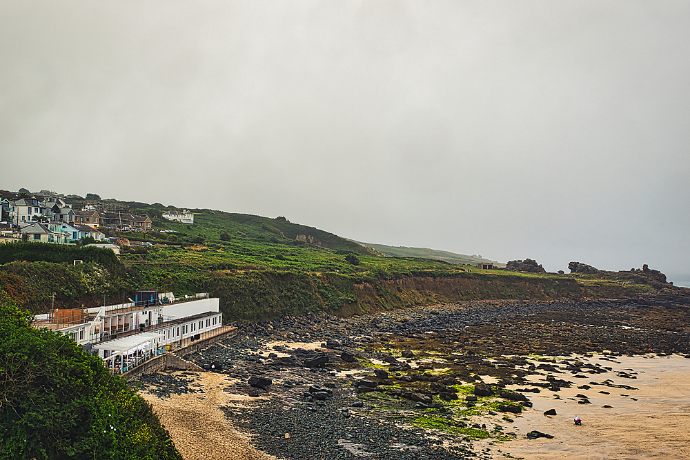 A coastal view featuring a rocky beach, a white building along the shore, and green hills in the background under a cloudy sky.