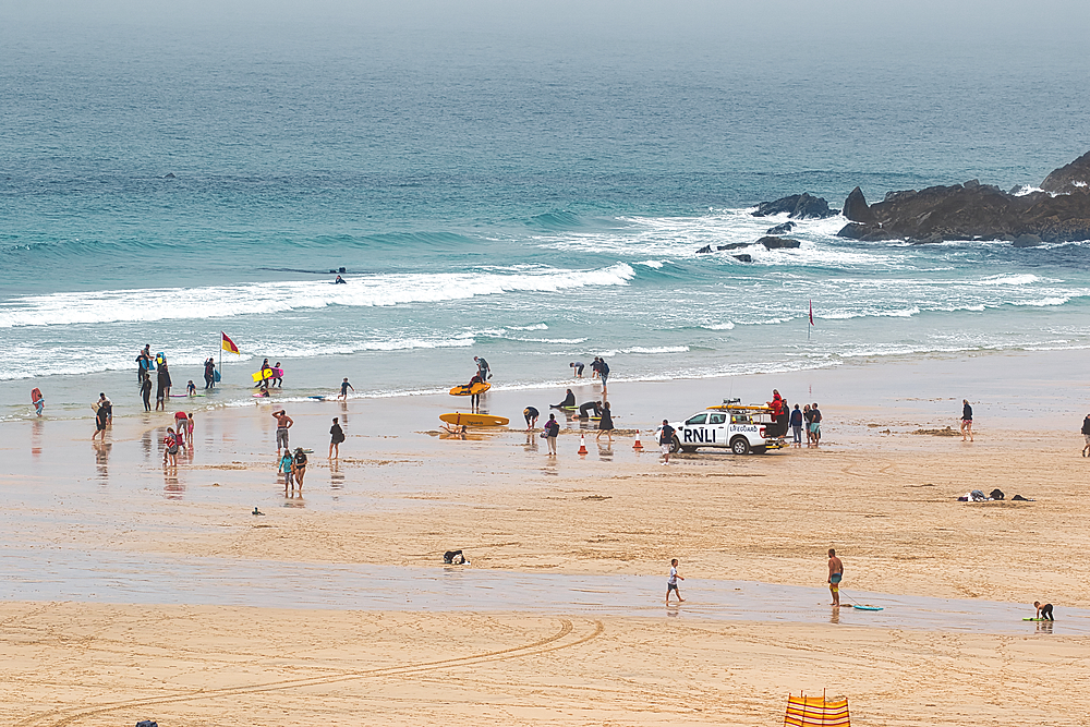 A busy beach scene with people enjoying various activities. Lifeguards are present, and some individuals are surfing in the water. The beach is sandy and wet, with a cloudy sky overhead.