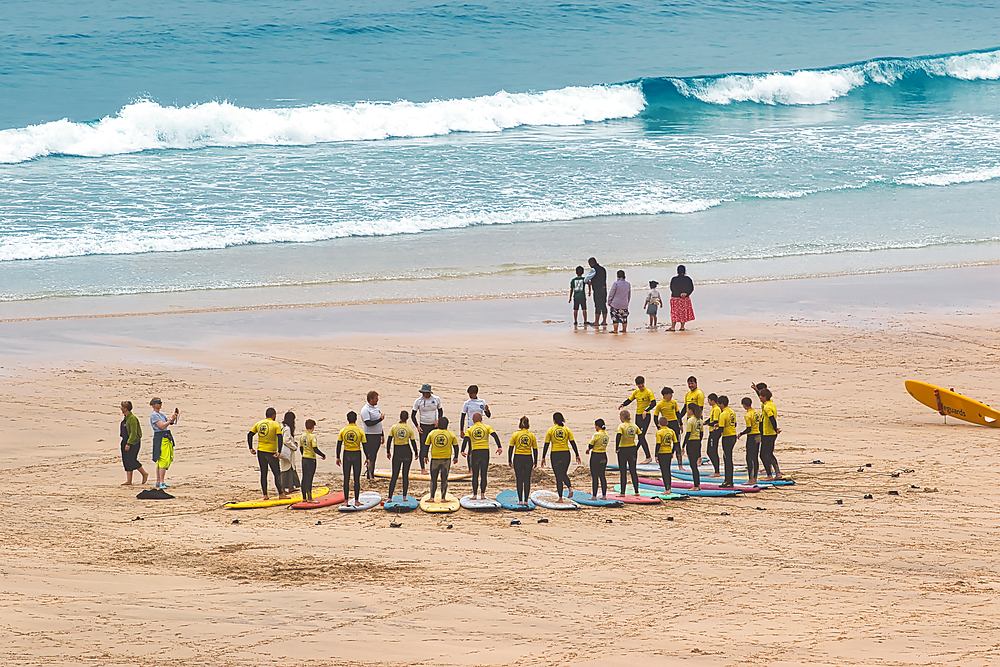 A group of surfers in yellow shirts gathered on the beach, preparing for a surfing lesson. The ocean waves crash in the background, and some people walk along the shore.