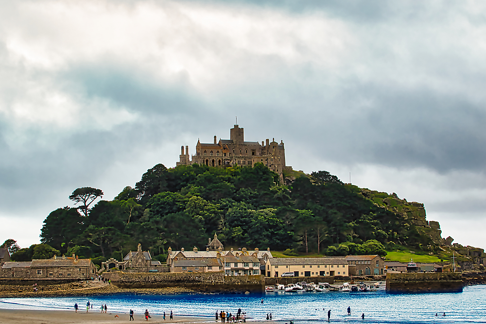 A scenic view of a castle atop a hill, surrounded by lush greenery and a cloudy sky. In the foreground, people stroll along a sandy beach, with boats moored in the water.