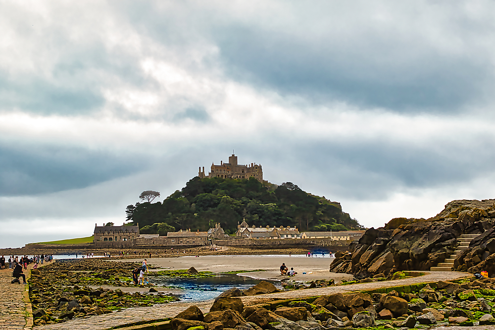 A scenic view of a coastal landscape featuring a castle on a hill, surrounded by rocky shores and a sandy beach. People are walking along the path and enjoying the scenery under a cloudy sky.