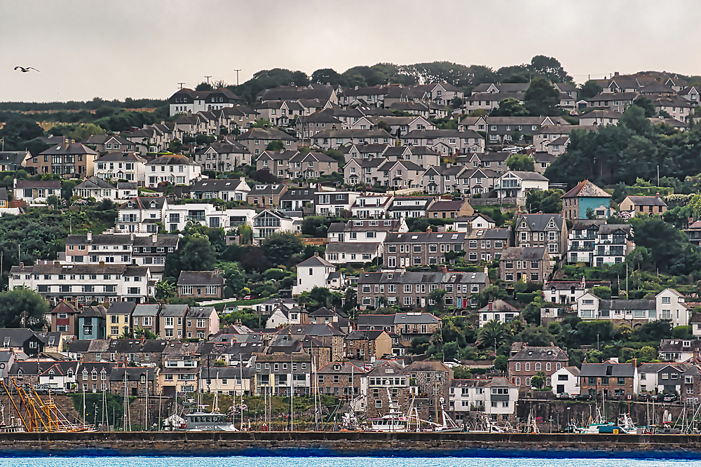 A scenic view of a coastal town with densely packed houses on a hillside, overlooking a harbor filled with boats. The sky is overcast, adding a moody atmosphere to the landscape.