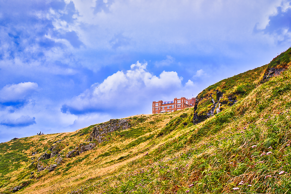 A scenic view of a hillside with lush green grass and rocky outcrops, featuring a distant building perched on the slope under a partly cloudy sky.