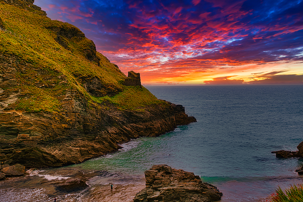 A stunning coastal landscape at sunset, featuring rocky cliffs and a vibrant sky filled with orange, pink, and purple hues. The calm sea reflects the colors of the sky, while a small figure stands on the beach.