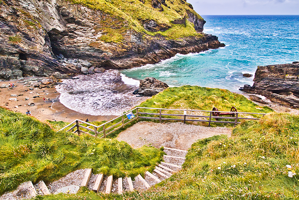 A scenic coastal view featuring a sandy beach surrounded by rocky cliffs and lush greenery. Steps lead down to the beach, with people enjoying the serene atmosphere.