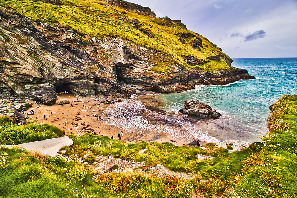 A scenic beach surrounded by rocky cliffs and lush greenery. People are enjoying the sandy shore and the gentle waves of the turquoise sea. The sky is partly cloudy, adding to the serene atmosphere.