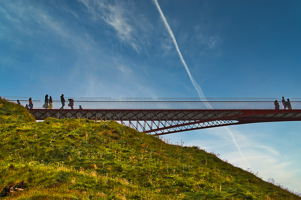 A silhouette of people walking on a bridge against a blue sky with wispy clouds. The bridge is elevated over a grassy hill, creating a scenic view.