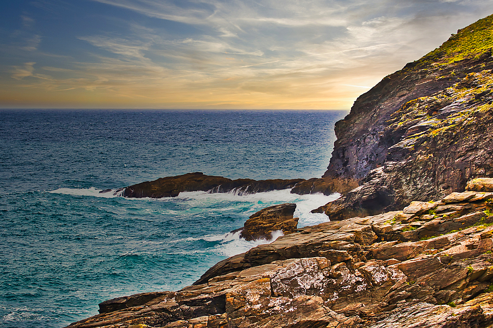 A scenic coastal view featuring rugged cliffs, crashing waves, and a serene sunset sky. The rocky shoreline contrasts with the deep blue ocean, creating a tranquil atmosphere.