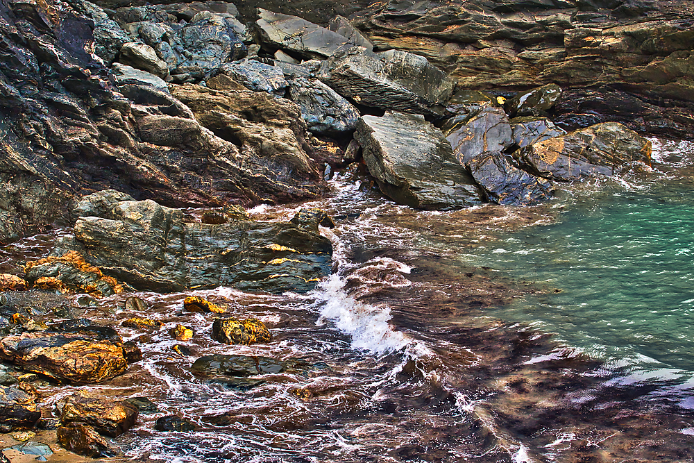 A rocky shoreline with waves gently crashing against the stones. The water is clear, revealing a mix of colors from the rocks and seaweed. The scene captures the natural beauty of the coast.