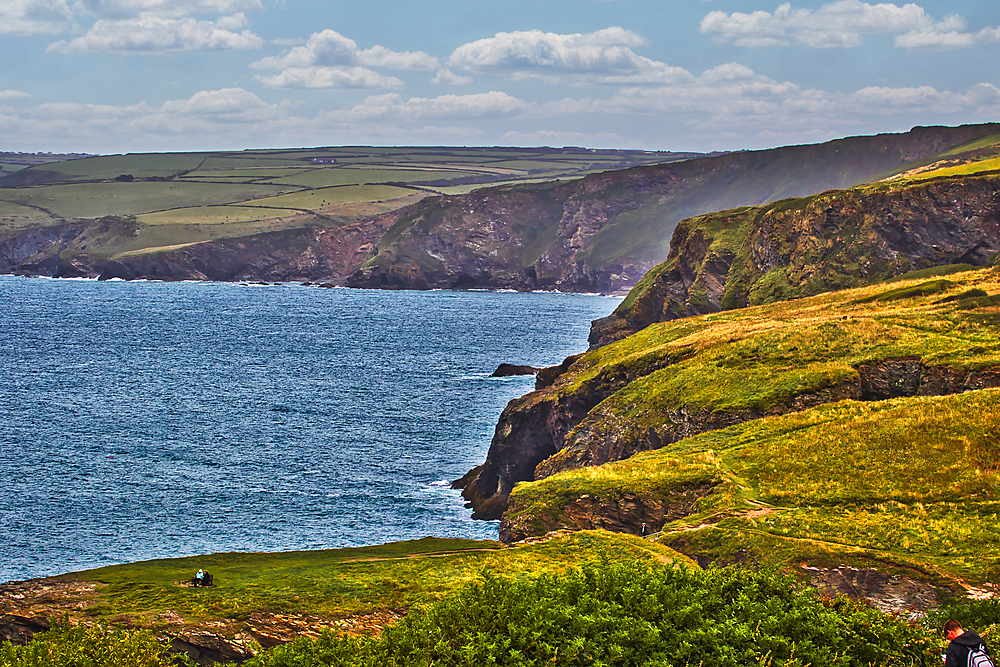 A scenic coastal view featuring rugged cliffs, lush green hills, and a calm blue sea under a partly cloudy sky. The landscape showcases the natural beauty of the coastline.