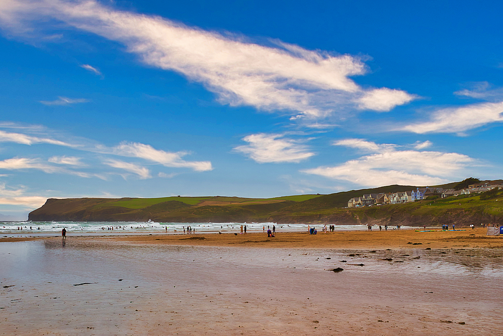 A scenic beach view with people enjoying the water, gentle waves, and a backdrop of green hills under a blue sky with wispy clouds.