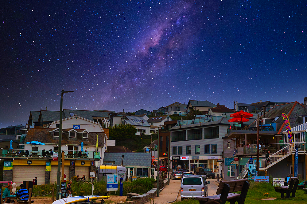 A picturesque coastal town at night with a starry sky and the Milky Way visible. The scene includes buildings, shops, and a beach area with vehicles parked.