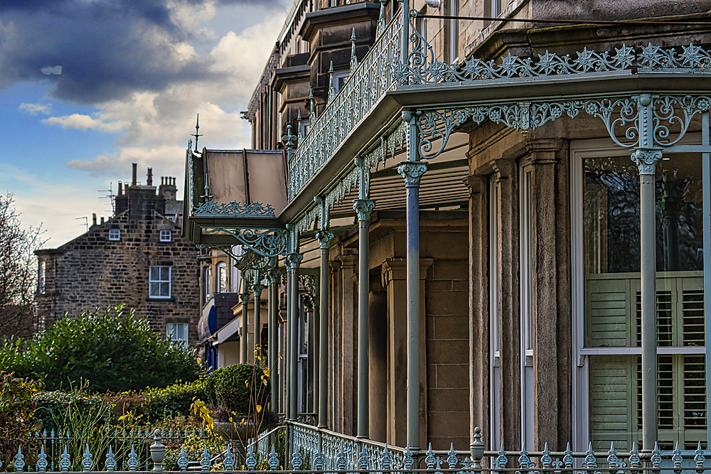 A row of Victorian-era terraced houses features ornate, light-green metalwork on its porches and railings. A stone house is visible in the background. The sky is partly cloudy.