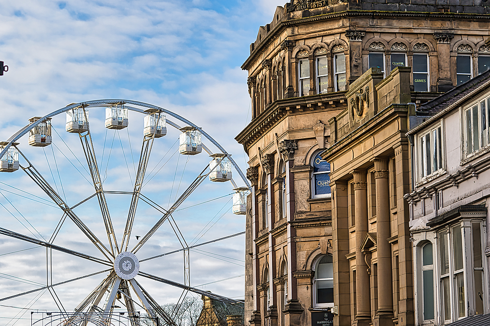 A Ferris wheel stands adjacent to a multi-story tan-colored building with classical architectural features. The sky is partly cloudy.