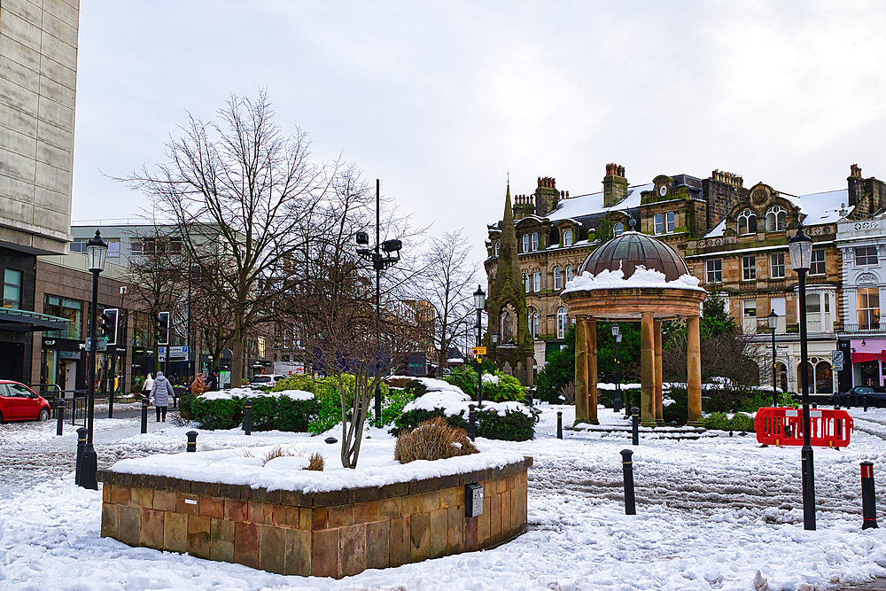 Snow-covered town square with a stone gazebo, buildings, and landscaping. Pedestrians are visible in the background in Harrogate, UK.