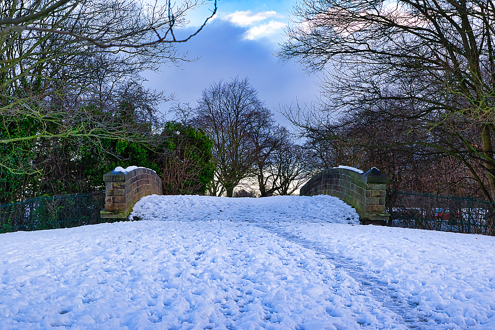 Snow-covered path leading to a stone low bridge between bare winter trees under a partly cloudy sky in Harrogate, UK.