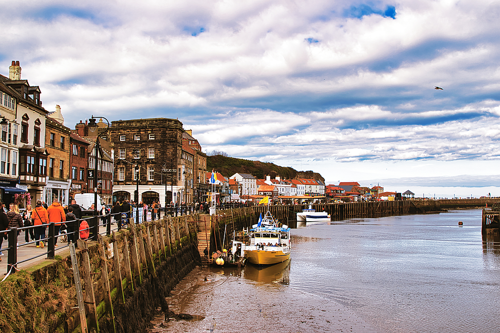 Scenic view of a coastal town featuring buildings, a harbor with boats, and a cloudy sky. People are walking along the waterfront in Whitby, UK.