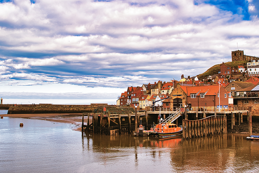 Coastal view featuring a harbor with a lifeboat, pier, distant lighthouse, and a hillside town under a cloudy sky in Whitby, UK.