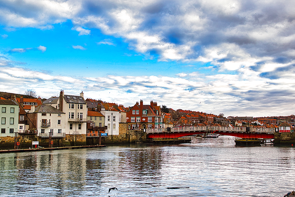 Waterfront view of town buildings along a river with a red bridge, under a partly cloudy sky reflecting in the water in Whitby, UK.