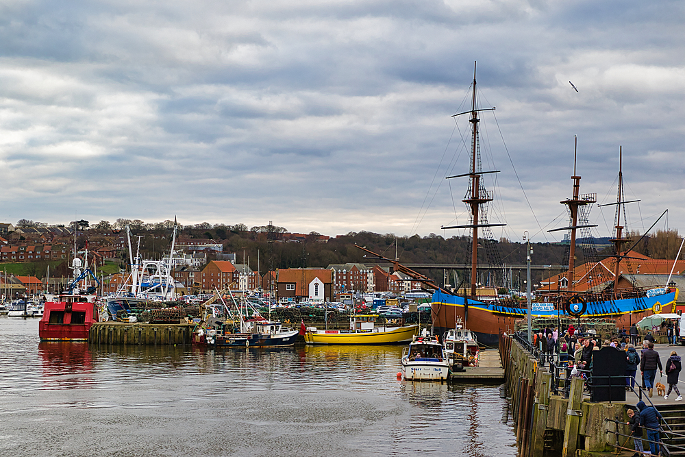 Harbor scene with various boats docked along a pier, buildings, and a cloudy sky. People are on the pier.