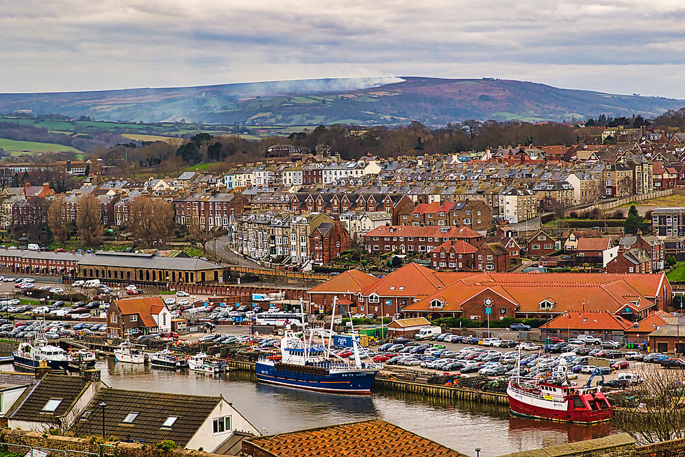 High angle view of a coastal town featuring buildings, a harbor with fishing boats, and a hillside under a cloudy sky in Whitby, UK.