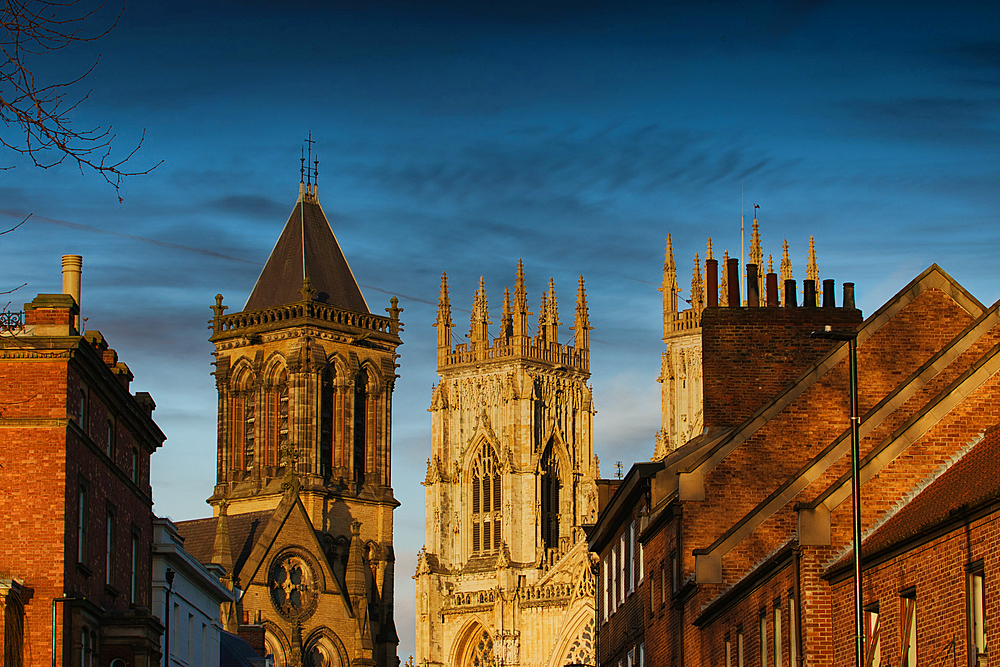 Image shows York Minster, England, flanked by brick buildings under a blue sky. The composition is eye-level, highlighting architectural details.