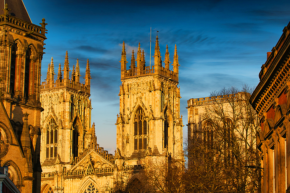 Exterior view of York Minster towers, with intricate Gothic details, under a blue sky. Warm sunlight illuminates the stonework.