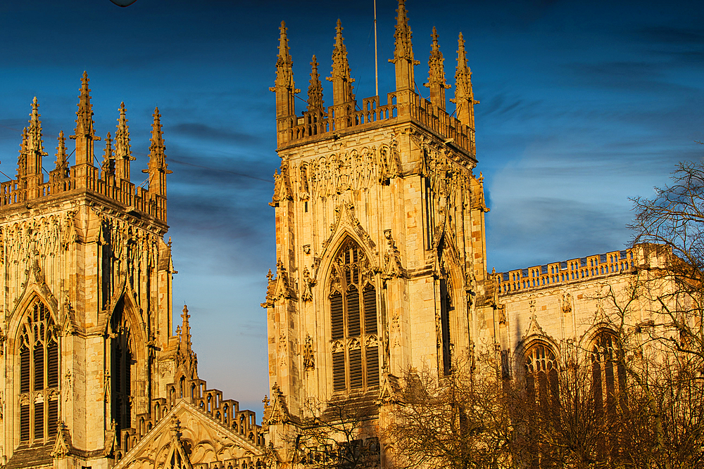 Two ornate, Gothic-style cathedral towers dominate the frame against a blue, slightly cloudy sky. Warm light illuminates the stone structures.