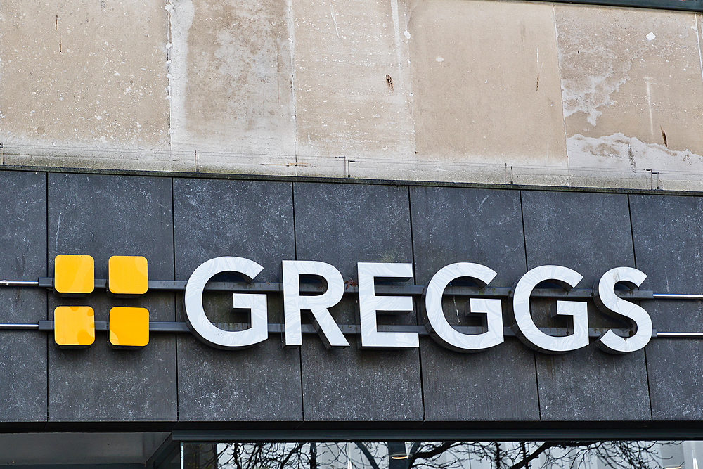 Greggs store sign. White lettering and orange squares on a dark gray tile background, with a lighter stone wall above in Liverpool, UK.
