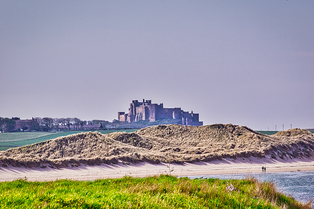 Landscape shot of a castle atop a hill, viewed from across grassy dunes, a sandy beach, and water. People are walking along the shoreline at Bamburgh Castle, UK.