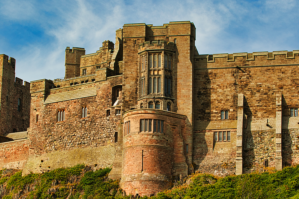 A large, imposing stone castle with multiple towers, walls, and windows, viewed from a low angle. Green vegetation at the base at Bamburgh Castle, UK.