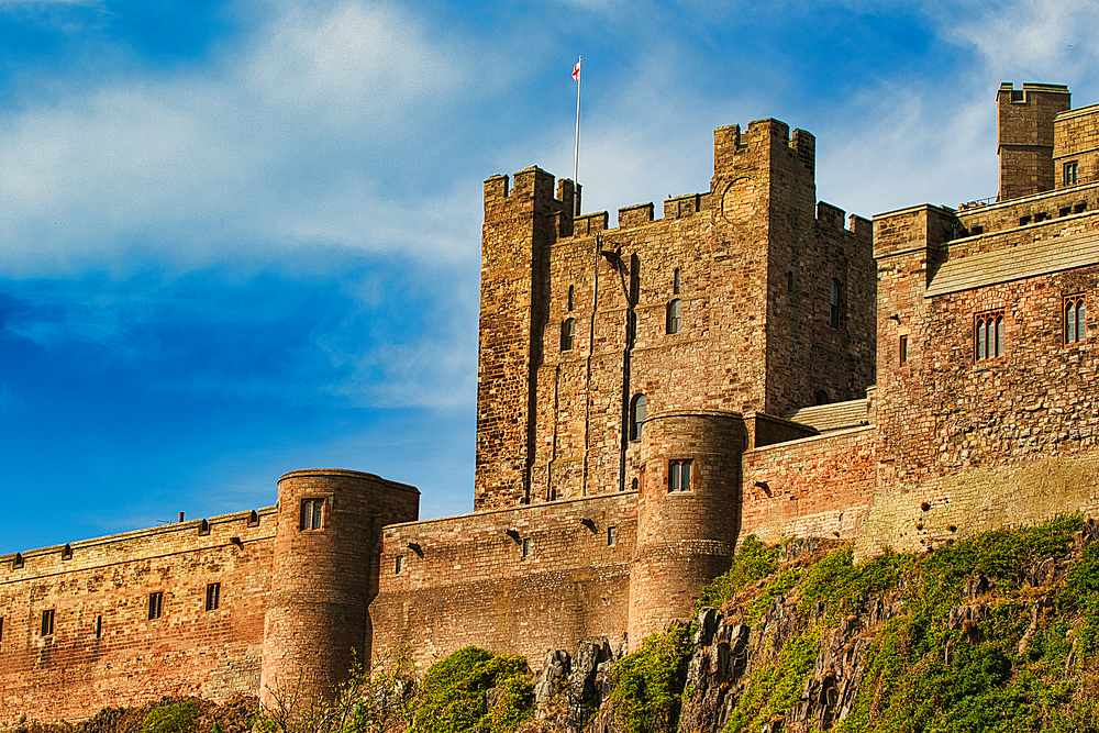 Low-angle view of a large, stone castle with turrets against a blue sky, situated on a rocky outcropping covered in greenery at Bamburgh Castle, UK.