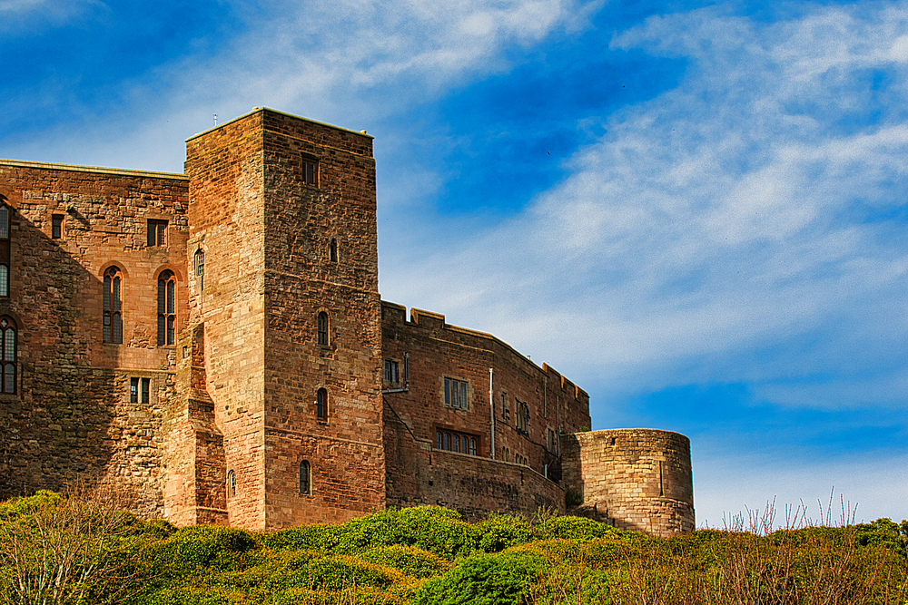A stone castle complex stands on a grassy hill against a blue sky with scattered white clouds at Bamburgh Castle, UK.