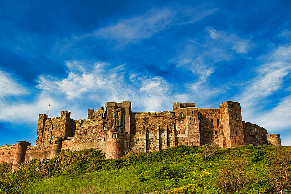 A wide shot of a stone castle on a grassy hill, set against a blue sky with scattered white clouds at Bamburgh Castle, UK.