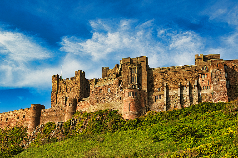 A large stone castle sits atop a green hill under a blue sky with wispy clouds. The castle is the dominant element at Bamburgh Castle, UK.