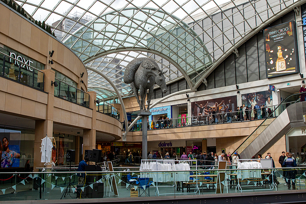Interior view of a multi-level shopping mall with a metal horse sculpture and various shops and people in Leeds, UK.
